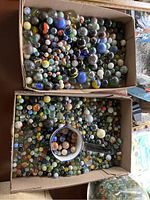 Two cardboard boxes filled with assorted glass and stone marbles and a ceramic mug with marbles inside