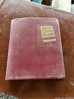 Closed maroon textured cover of the vintage match cover album showing gold and black lettering.