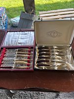 Two open boxes on a table showing two complete sets of six vintage steak knives each with serrated blades and tortoiseshell-style handles.