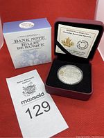 Photo of the silver coin in its red display case with the original box behind and auction tag.