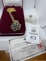 Gold-tone long necklace with blue floral pendant on burgundy velvet pad inside jewelry box. Certificate of authenticity visible.