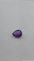 Close-up image of a deep purple, pear-shaped amethyst gemstone on a white background showing its size and cut.