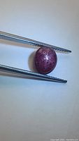 Close-up image of an oval cabochon cut red ruby held by tweezers on a plain background, showing surface texture and star effect.