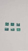 Six rectangular baguette cut Colombian emerald gemstones arranged on a white background showing color and clarity.