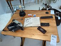 Wood table displaying Kodak Tourist camera with open bellows, Edison filament lamp, two pairs of glasses, open book/manual, two small boxes, leather case for razor kit, and matchbook.
