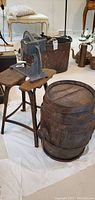 Photo showing antique metal fruit press mounted on a rustic wooden stool and nearby antique wooden wine barrel with metal bands on the floor.
