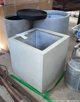 Three composite planters in varying size and a black ceramic bowl placed on top of one planter, shown in garage setting with surrounding tools and watering can.