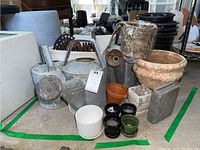 Overview photo showing multiple watering cans, ceramic and plastic planters, garden edger, and antique tool arranged together on ground.