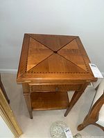 Front view of wooden fold top game table closed showing geometric inlay top, drawer, and lower shelf.
