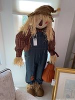 Front view of large stuffed scarecrow figure standing, showing face, clothing, and pumpkin accessory.