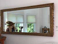 Front-facing photo showing the large rectangular vintage wall mirror with an ornate golden enameled wood frame hanging on a white wall. Reflection visible, including window blinds and décor.