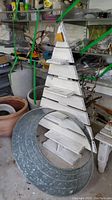 Photo showing rustic wooden pine tree-shaped display shelf and galvanized metal tree collar on the floor in garage-like setting with storage items and pots behind.