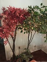 Photo showing both faux trees side by side, one with red maple leaves and the other with green foliage, both with string lights and wicker baskets at bases.