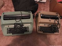 Photo showing two vintage IBM Selectric electric typewriters side by side on carpeted floor, whitish beige machine on the right and green machine on the left with cords coiled over the keyboards.