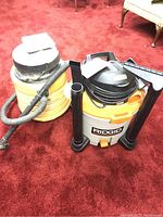 Photo shows both wet vacuums side by side on red carpet, RIDGID wet vac on right with hose and attachments, Genie wet vac on left with silver top.