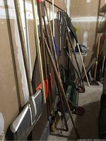 Photo of assorted garden tools including metal rakes, shovels, and other yard equipment arranged against a wall in a garage, showing some rust and wear.