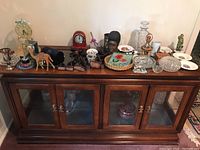 Photo showing various decorative items including non-working glass-domed anniversary clock, smaller red mantle clock, carved wood figurines, glassware and collectibles arranged on the wooden cabinet.