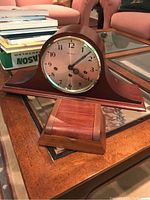 Full view of wood mantle clock on wood table with glass top, curved base and round clock face, showing overall clock design and finish.