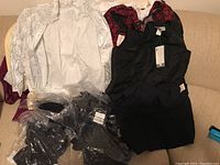 Photo showing white patterned blazer, black sleeveless top, red and black dress, and other tops folded on couch.