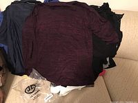 Pile of women's tops in dark colors, mostly long sleeves, and what appears to be plastic packaging bags on a beige sofa.