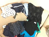 Photo showing multiple vintage women's clothing items including a black floral dress, navy dress with blue stripes, light blue blouse/jacket, white polka dot blouse, and other items loosely arranged on a beige couch.