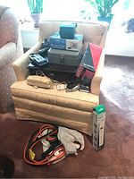 Overview of various office supplies and tools placed on a beige chair and surrounding floor including boxes, vintage telephone, car jumper cables and a boxed fluorescent light bulb.