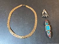 Three pieces sterling jewelry arranged on a dark surface featuring a gold-tone four strand braided necklace, a love bird pin, and a Southwest style watch with turquoise and coral stones.