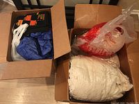 Two cardboard boxes filled with various vintage linens and miscellaneous items including Halloween-themed fabrics and white linens packed in plastic