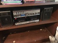 Full view of Panasonic stereo system RX-CW50 showing main unit with dual cassette decks and two bookshelf speakers on either side. Dust and debris visible on shelf surface.