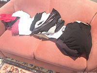 Multiple tops, jackets, and dresses layered on a sofa, showing variety of colors and styles including red, white, black, and patterned pieces.