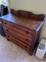 Full view of the antique chest of drawers showing three drawers with wooden pulls, decorative curved backboard, and worn top surface.
