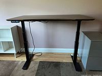 Full front view of electric standing desk with digital height control panel visible on left side, placed on wood floor between two shelves.