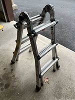 Aluminum folding step ladder partially collapsed inside an open garage with concrete floor and pavement outside. Ladder has black plastic hinge joints and foot grips.