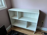 Photo showing white Ikea bookcase positioned against light purple wall near window, with four open shelves, modern minimalist design, and wood grain texture.