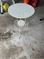 White Ikea metal side table with round top, three curved legs, and small circular bottom shelf shown on concrete floor with some stains.