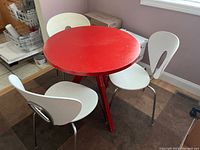 Red round table with three angled legs and four white modern plastic chairs with metal frames, showing table surface wear