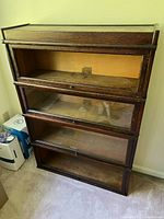 Front view of four-tier wooden physician's bookcase with glass doors showing missing glass pane on top shelf and wear marks on wood surface.