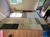 Full view of the rectangular wool rug laid on a wooden floor beside windows and door, showing geometric patchwork pattern with various shades of green and black blocks