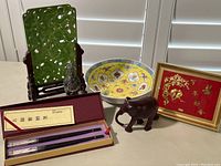 Full view of the lot showing the faux jade carving on stand, decorative bowl, carved wood elephant, framed gold-tone artwork, stone figure, and boxed chopsticks.