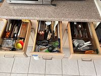 Three kitchen drawers open showing kitchen utensils and flatware including spoons, forks, knives, chopsticks, scissors, ladles and measuring cups.
