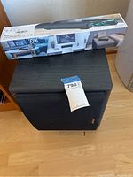 Photo showing black fabric-covered subwoofer with tagging label on top, standing on wooden floor.
