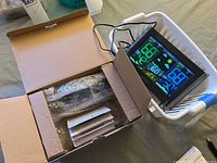 Open box with wrapped clock and instruction manuals inside, plus separate digital weather monitor displaying temperature and humidity.