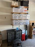 Photo showing shelves with fabric storage bins in white and pale blue with flap tops, clear plastic shoebox containers, and small wooden compartment organizer on top shelf, plus two black plastic drawer units below.