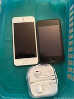 Top-down view of white and black iPod Touch devices alongside white Apple earphones in plastic case.