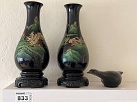Two matching decorative vases with green mountain and golden tree design on a black background, each approximately 11 inches tall, displayed on black carved pedestals; solid metal bird sculpture with a matte finish next to them.