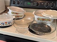 Full lot displayed on stove top with four Corningware dishes, three with glass lids, two Pyrex pie plates, and one clear glass heart-shaped dish.