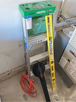 Full lot with Werner ladder green top, yellow construction level, Tiro Power Sweep leaf blower black unit, and coiled orange extension cord on floor by wall
