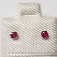 Front view of earrings showing the small round red rubies in four-prong 14K gold settings on a white padded display card.