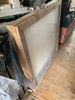 Side angled view showing the solid wood frame with glass front and fiberboard backing, surface shows signs of dust and storage wear.