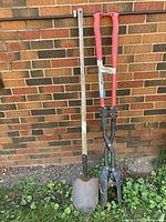 Shovel and fence post digger leaning against brick wall, showing full length and condition of each tool.
